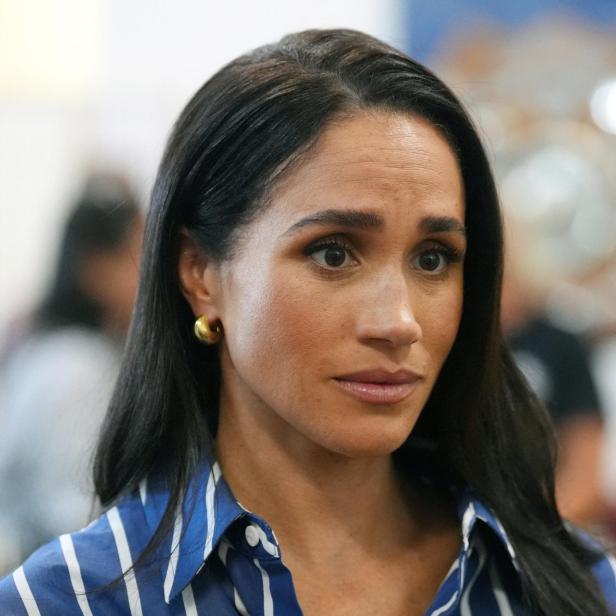 Meghan Markle trägt ein blau-weiß gestreiftes Hemd und blickt mit ernstem Ausdruck zur Seite.