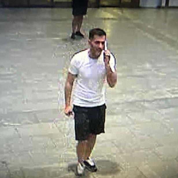 Ein Mann in weißem T-Shirt und schwarzer Shorts spricht über ein Handy und geht auf einem Platz.