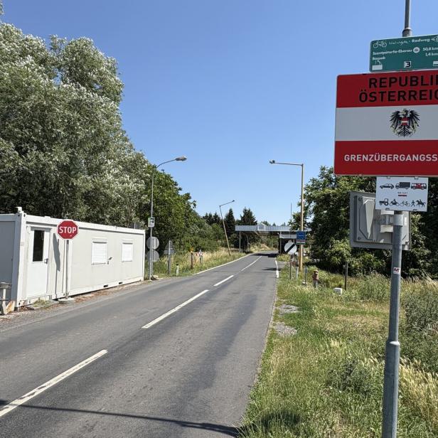 Grenzübergang nach Österreich mit Straßenschild, Kontrollhäuschen und leerer Landstraße unter klarem Himmel.