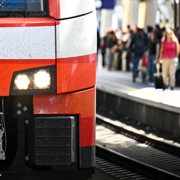 Die Front eines rot-weißen ÖBB-Zuges von Siemens steht an einem Bahnsteig, auf dem unscharf erkennbare Menschen warten.