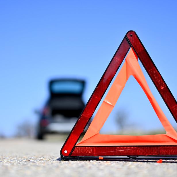 Ein rotes Warndreieck steht auf einer Straße vor einem Auto mit geöffnetem Kofferraum.