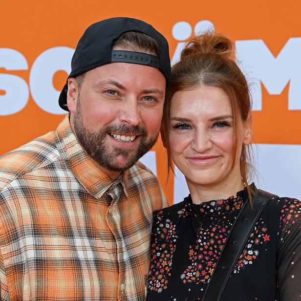 Felix von Jascheroff lächelt neben seiner Ehefrau Alexandra-Sophie Winterberg vor einer orangen GZSZ-Medienwand.