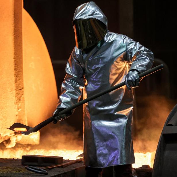 Schweißer in Hitzeschutzkleidung arbeitet mit einer langen Stange vor glühendem Metall und Feuer.