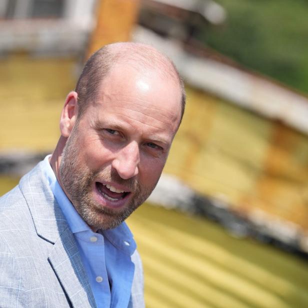 Prinz William trägt ein hellblaues Hemd und ein graues Sakko vor einem unscharfen, gelblichen Hintergrund.