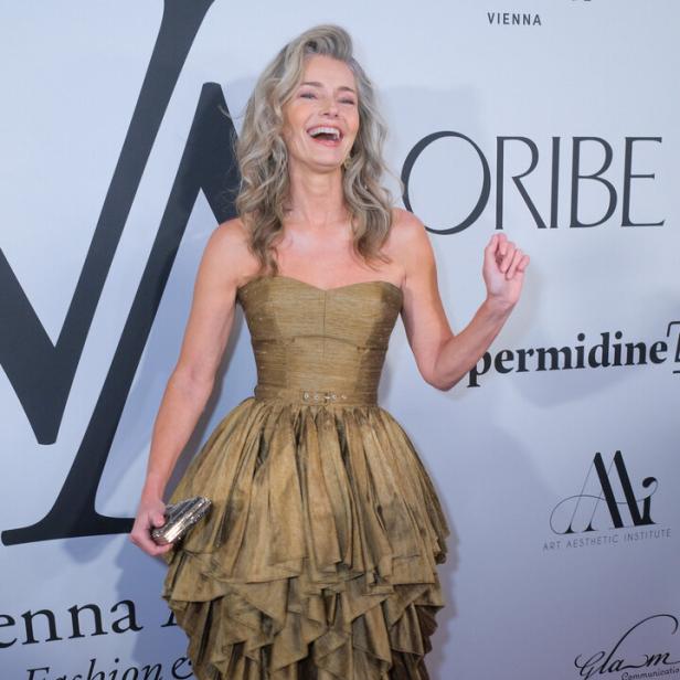Paulina Porizkova mit grauem Haar lacht und trägt ein trägerloses, goldfarbenes Kleid mit Stufenrock vor einer Sponsorenwand.