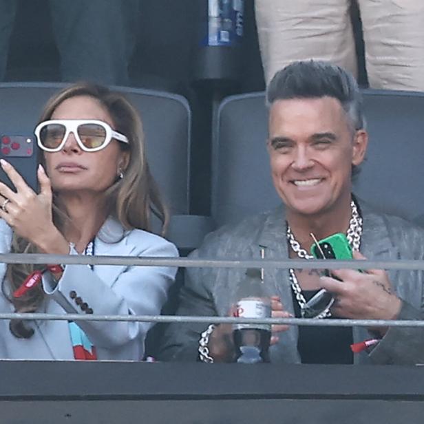 Ayda Field und Robbie Williams sitzen lächelnd auf der Tribüne, während Ayda ein Foto mit ihrem Smartphone aufnimmt.