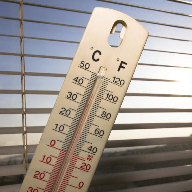 Ein Thermometer zeigt vor einem sonnigen Fenster mit Jalousien eine Temperatur von etwa 35 Grad Celsius an.