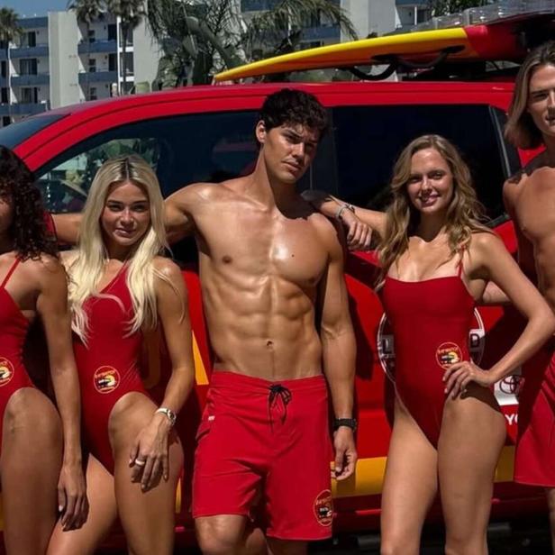 Fünf Rettungsschwimmer in roten Badeanzügen und Shorts posieren vor einem roten Lifeguard-Pick-up mit Surfbrettern auf dem Dach.