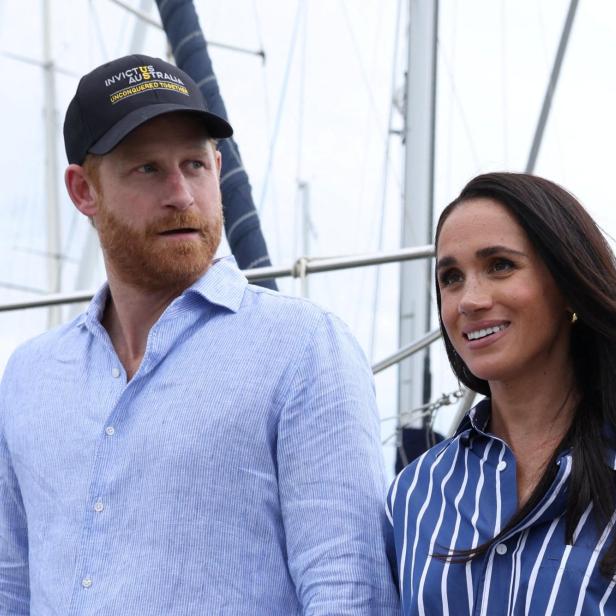 Prinz Harry mit einer Invictus-Games-Kappe und Meghan Markle stehen vor Segelbooten.