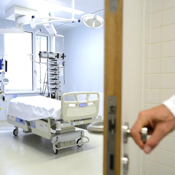Operationssaal mit leerem Krankenhausbett und medizinischen Geräten, eine Hand öffnet die Tür.