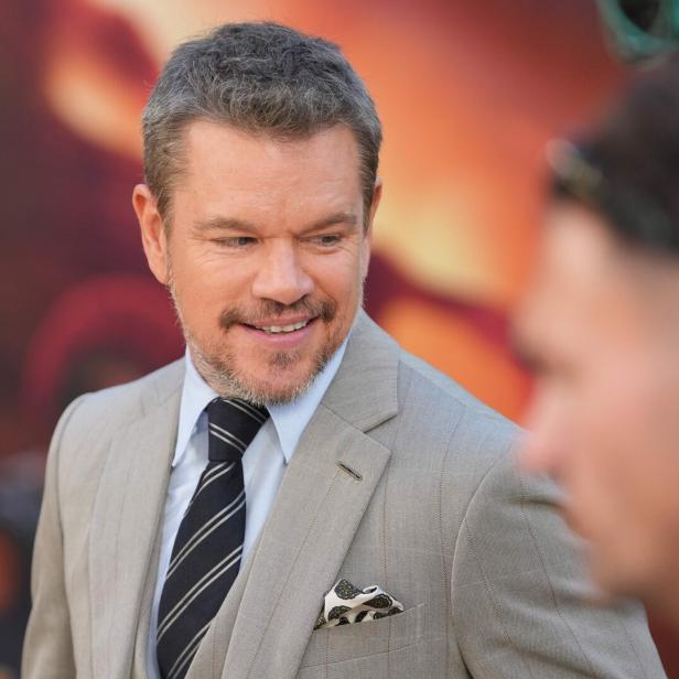 Matt Damon lächelt in einem hellen Anzug mit Krawatte vor einem unscharfen, orangefarbenen Hintergrund.