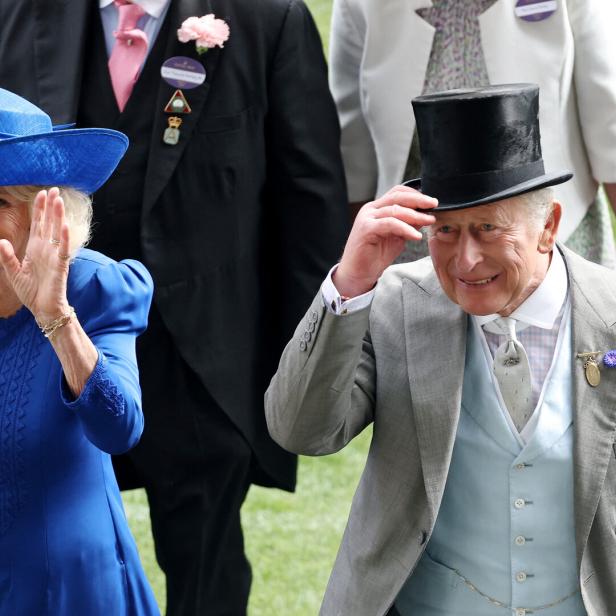 König Charles III. und Königin Camilla winken bei einem öffentlichen Auftritt, wobei Charles seinen Zylinder leicht anhebt.