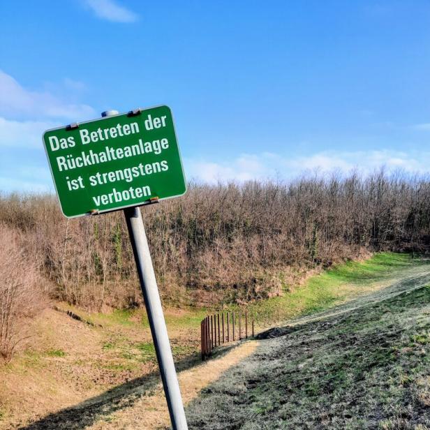 Grünes Verbotsschild an einem Rückhaltebecken vor hügeliger Wiese und blauem Himmel.