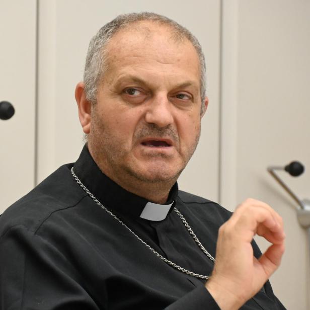 Der syrische Erzbischof Jacques Mourad sitzt in schwarzem Priestergewand vor einer weißen Wand.