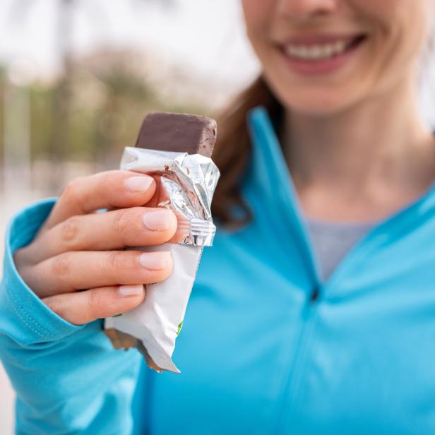 Lächelnde Frau in blauer Jacke hält einen ausgepackten Schokoriegel in der Hand.