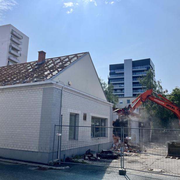 Bagger reißt ein Gebäude neben einem weißen Haus ab.
