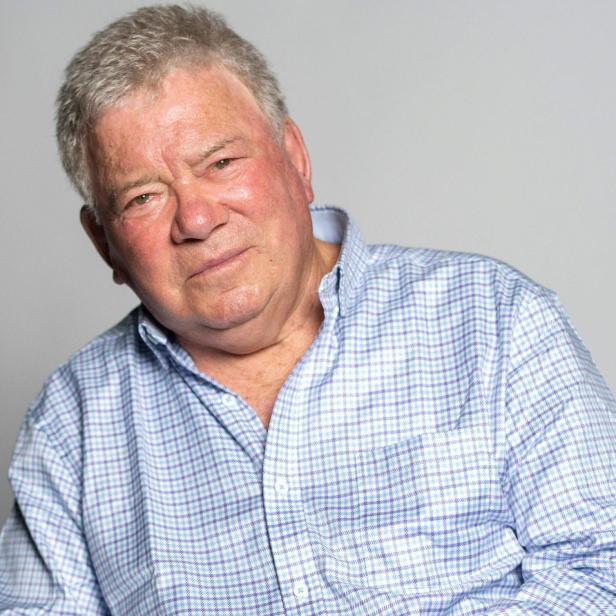 William Shatner trägt ein hellblau kariertes Hemd und blickt vor einem neutralen Hintergrund in die Kamera.