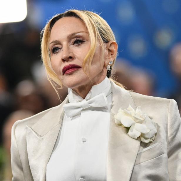 Madonna trägt einen hellen Anzug mit weißer Fliege und einer weißen Blume am Revers vor einem unscharfen Hintergrund.
