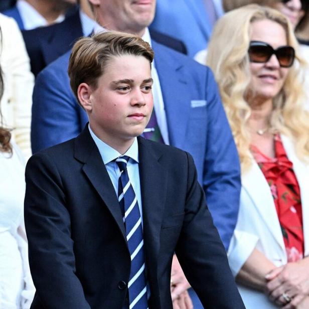 Prinz George trägt einen dunklen Anzug mit Krawatte und stützt sich auf eine hölzerne Brüstung.