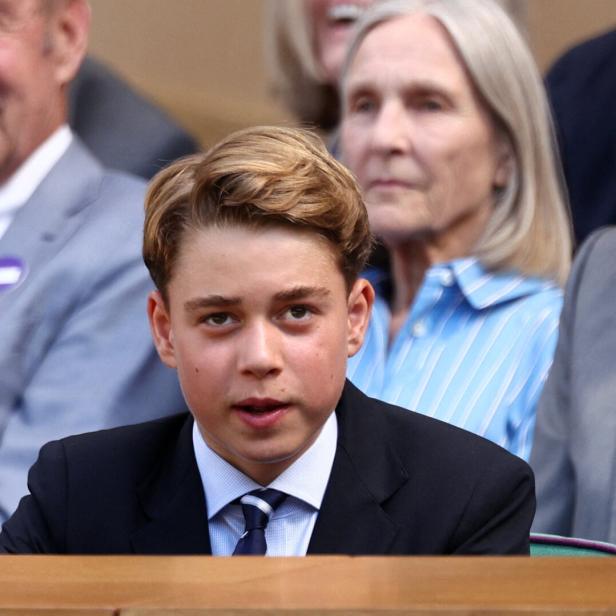 Prinz George sitzt in einem dunklen Anzug mit Krawatte auf der Tribüne, während im Hintergrund weitere Personen zu sehen sind.