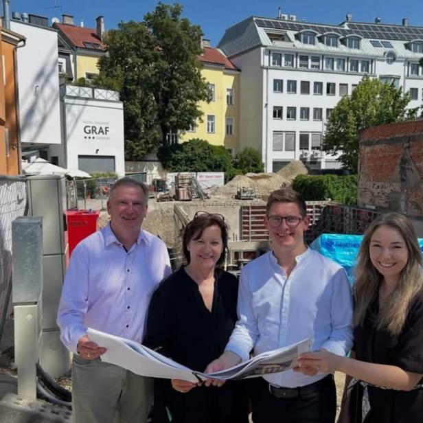Vier Personen stehen vor einer Baustelle und halten gemeinsam Pläne.