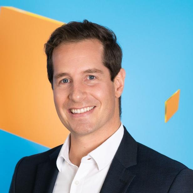 Geschäftsführer Philipp König lächelt, im Anzug vor blauem Hintergrund mit orangefarbenen Flächen, über gute Zahlen der kronehit-Senderfamilie.