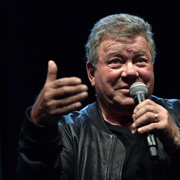 William Shatner spricht vor einem dunklen Hintergrund in ein Mikrofon und gestikuliert mit der rechten Hand.
