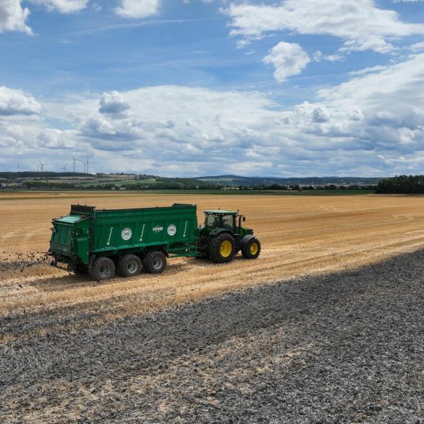 Traktor mit grünem Anhänger fährt über ein abgeerntetes Feld unter wolkigem Himmel.