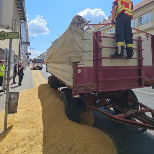 Ein Traktor mit Anhänger hat Maiskörner auf die Straße verloren, während ein Feuerwehrmann darauf steht.