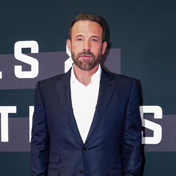 Ben Affleck trägt einen dunkelblauen Anzug mit weißem Hemd vor einer dunklen Hintergrundwand mit Schriftzügen.