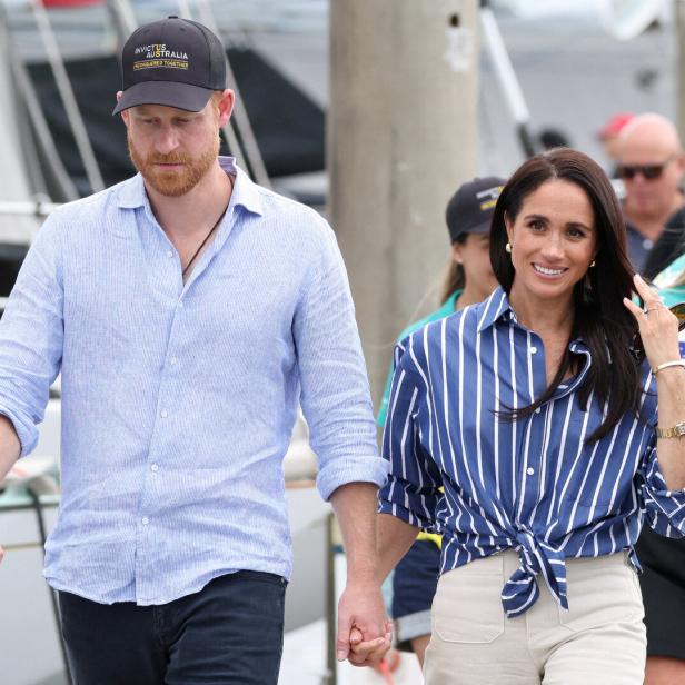 Prinz Harry und Herzogin Meghan gehen händehaltend an einem Hafen entlang, wobei Harry eine Invictus-Australia-Kappe trägt.
