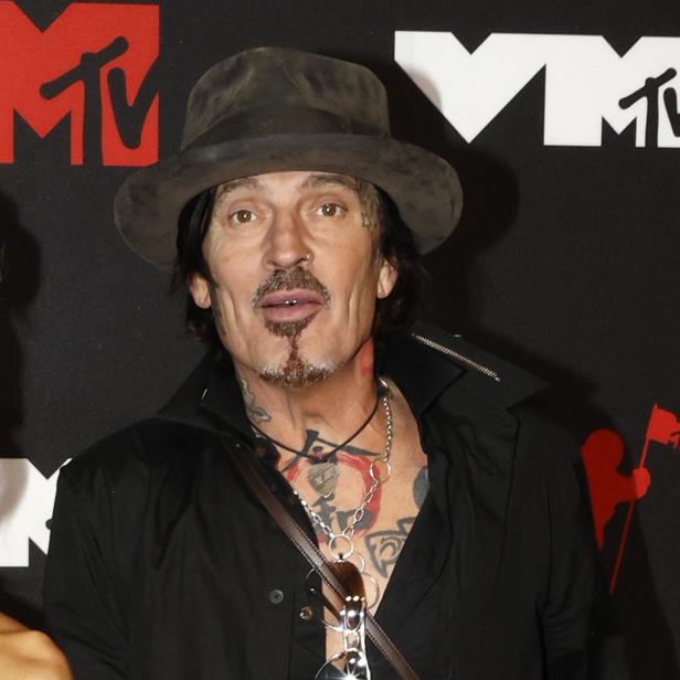 Tommy Lee trägt einen Hut und ein schwarzes Hemd vor einer MTV-Hintergrundwand.