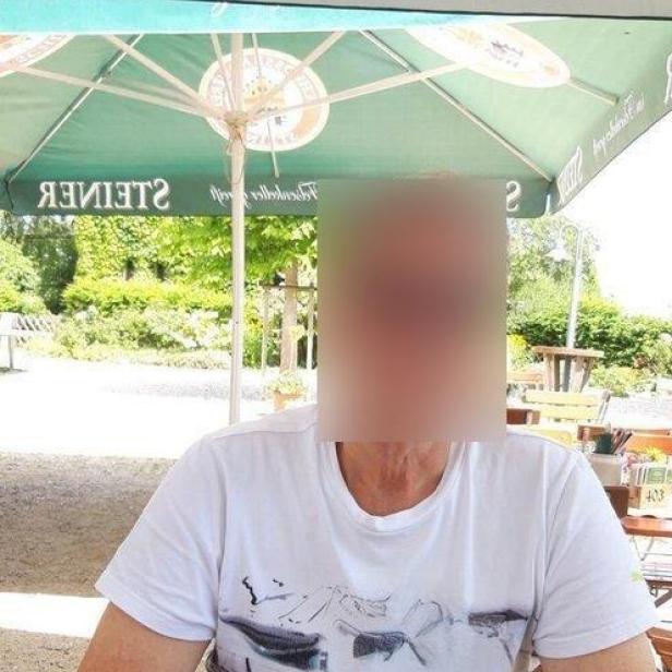 Eine Person sitzt im Freien an einem Tisch unter einem großen Sonnenschirm; das Gesicht ist unkenntlich gemacht.