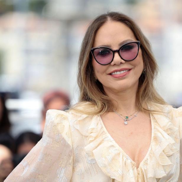 Ornella Muti lächelt in einem hellen, gerüschten Kleid und mit einer getönten Sonnenbrille vor einer unscharfen Kulisse.