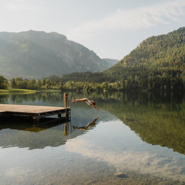 Eine Frau springt von einem Steg in einen stillen Bergsee, umgeben von bewaldeten Hängen und Spiegelungen im Wasser.