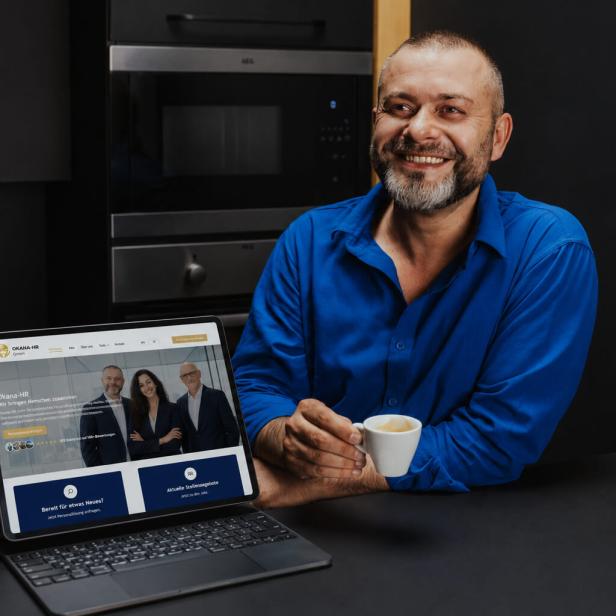Mann mit Bart und blauem Hemd sitzt an schwarzem Tisch und hält weiße Tasse neben offenem Laptop mit Webseite.