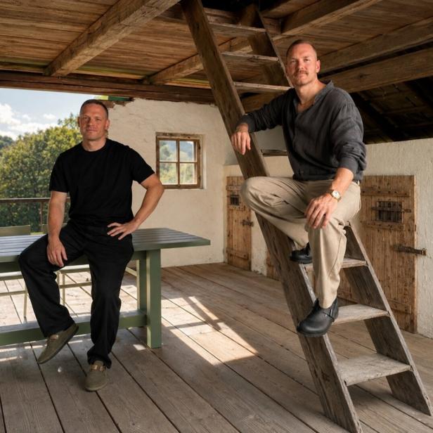 Zwei Männer posieren auf einer rustikalen Holzterrasse, einer sitzt am Tisch, der andere auf einer Leiter.
