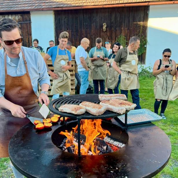 Mann grillt Fleisch und Gemüse an einer offenen Feuerstelle, während mehrere Personen in Schürzen zuschauen.