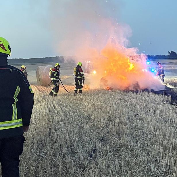 Feuerwehrleute löschen einen brennenden Wagen auf einem Feld, während Rauch und Flammen aufsteigen.
