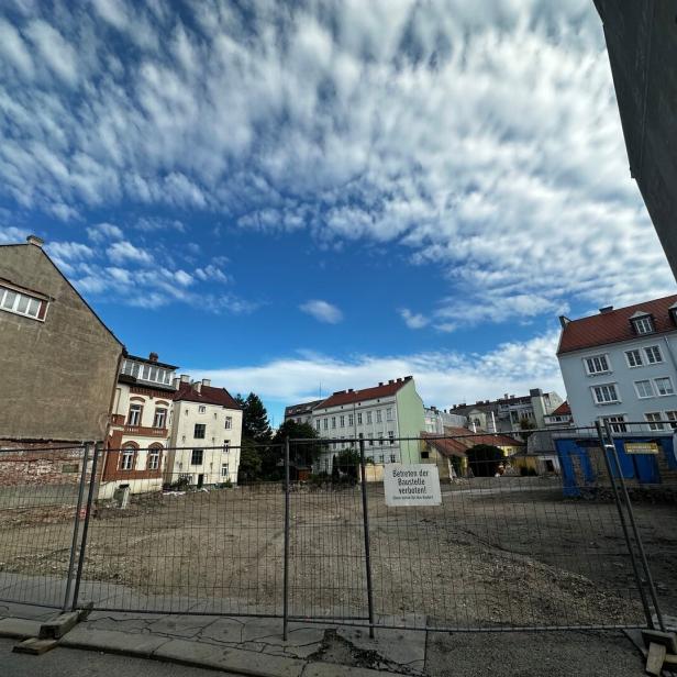 Umzäunte Baustelle zwischen Wohnhäusern unter dramatischem Wolkenhimmel.