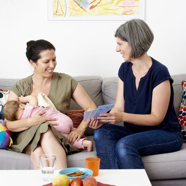 Eine Frau mit einem Säugling sitzt auf einem Sofa, daneben sitzt eine Familienbegleiterin, die ihr Informationsmaterial zeigt.