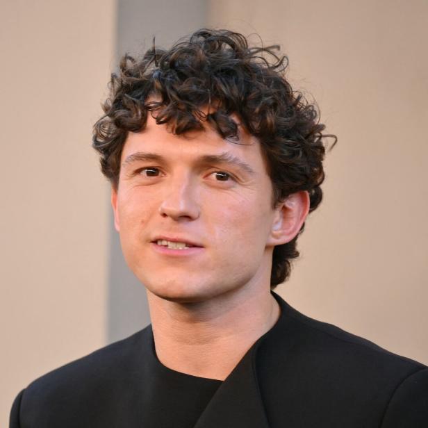 Tom Holland mit lockigem Haar und schwarzem Oberteil vor einem neutralen Hintergrund.