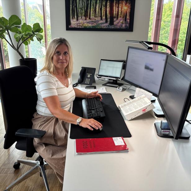 Frau sitzt an einem modernen Schreibtisch im Büro mit zwei Monitoren und Tastatur, umgeben von Pflanzen und Tageslicht.