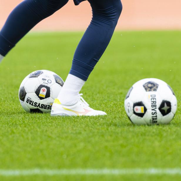 Fußballspiel mit zwei Bällen und den Beinen einer Spielerin auf dem Rasen.
