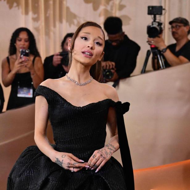Ariana Grande trägt ein schwarzes schulterfreies Kleid und eine Halskette, während sie von Fotografen umgeben ist.