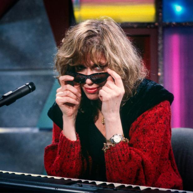 Carly Simon sitzt am Klavier und zieht ihre Sonnenbrille zurecht.