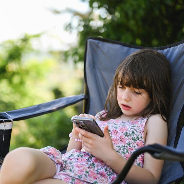 Ein Mädchen sitzt in einem Campinghocker und tippst auf einem Smartphone.