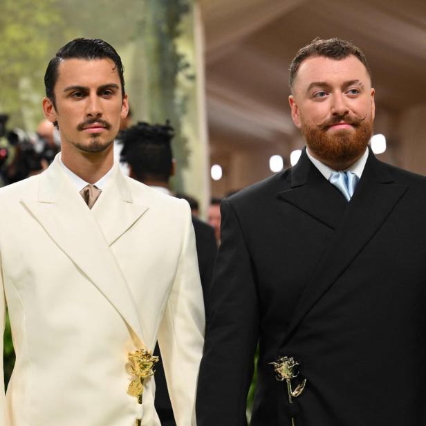 Sam Smith und Christian Cowan in eleganten Anzügen mit goldenen Rosen-Anstecknadeln bei einer Veranstaltung.