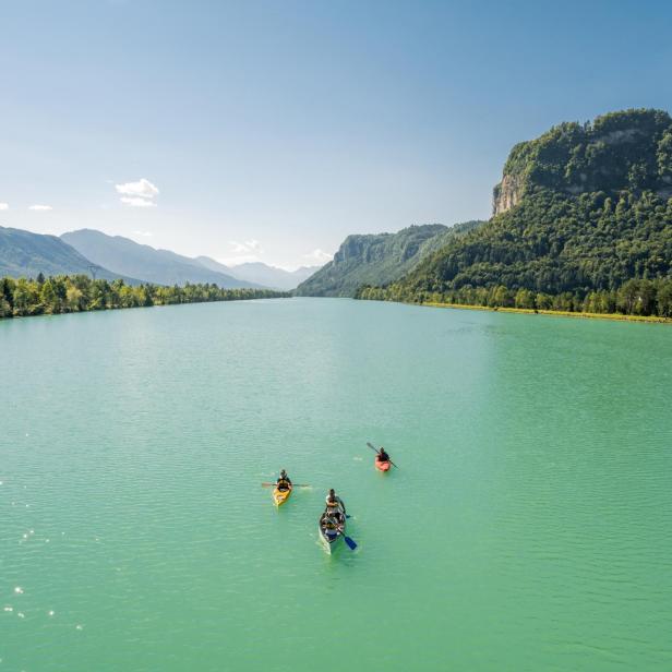 Drei Boote mit Menschen paddeln auf einem türkisfarbenen Fluss vor einer bewaldeten Bergkulisse.