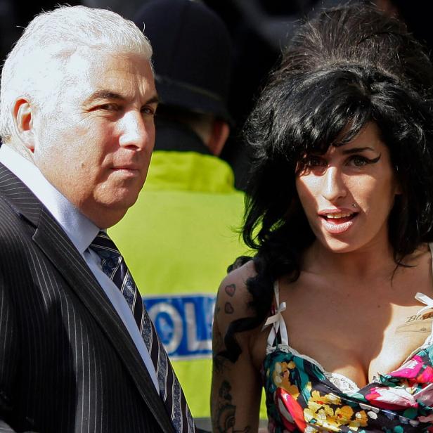 Amy Winehouse steht neben ihrem Vater Mitch im Anzug, während im Hintergrund ein Polizist in neongelber Weste zu sehen ist.
